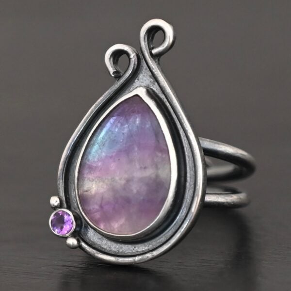 Close up of the "Elliana" Ring. Made out of sterling silver and fluorite and amethyst. The fluorite is the main stone in a teardrop shape. There is an amethyst in the bottom left corner of the ring. The silver comes up to the top to create two loops that are connected, one is taller than the other. The ring is resting against a dark background.