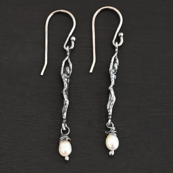 Close up of the "Goddess" Earrings. These earrings are made out of sterling silver and pearls with french hook ear wires. The earrings are sterling silver "women". The pearl hangs from the bottom of the earrings. The pearl is wrapped with sterling silver that is shiny. The rest of the sterling silver is oxidized. Earrings are shown against a dark background.
