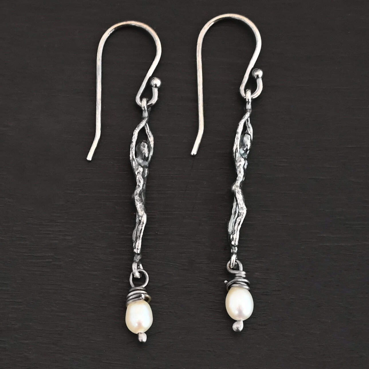 Close up of the "Goddess" Earrings. These earrings are made out of sterling silver and pearls with french hook ear wires. The earrings are sterling silver "women". The pearl hangs from the bottom of the earrings. The pearl is wrapped with sterling silver that is shiny. The rest of the sterling silver is oxidized. Earrings are shown against a dark background.
