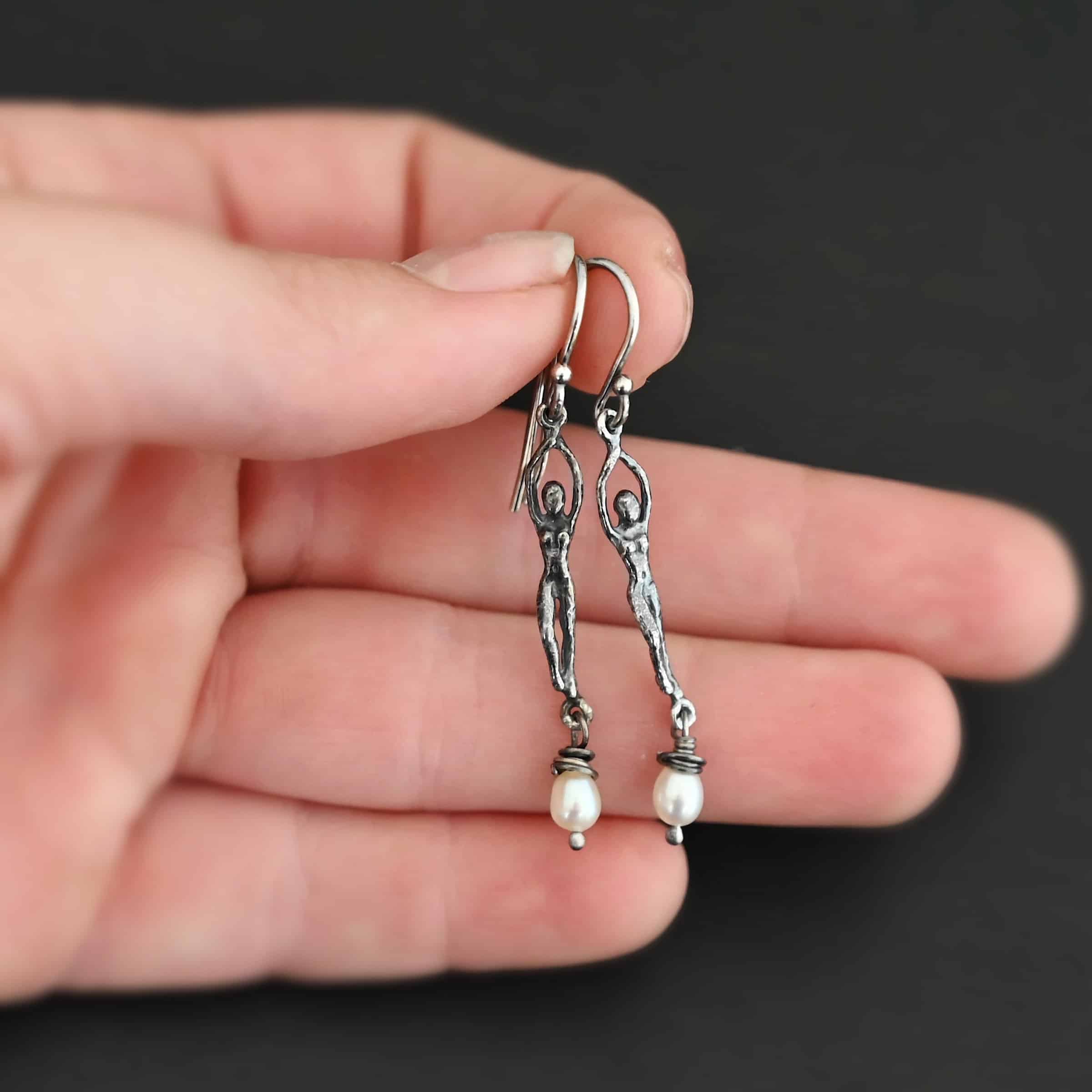 Close up of the "Goddess" Earrings. These earrings are made out of sterling silver and pearls with french hook ear wires. The earrings are sterling silver "women". The pearl hangs from the bottom of the earrings. The pearl is wrapped with sterling silver that is shiny. The rest of the sterling silver is oxidized. Earrings are shown on the palm of a hand to demonstrate size.