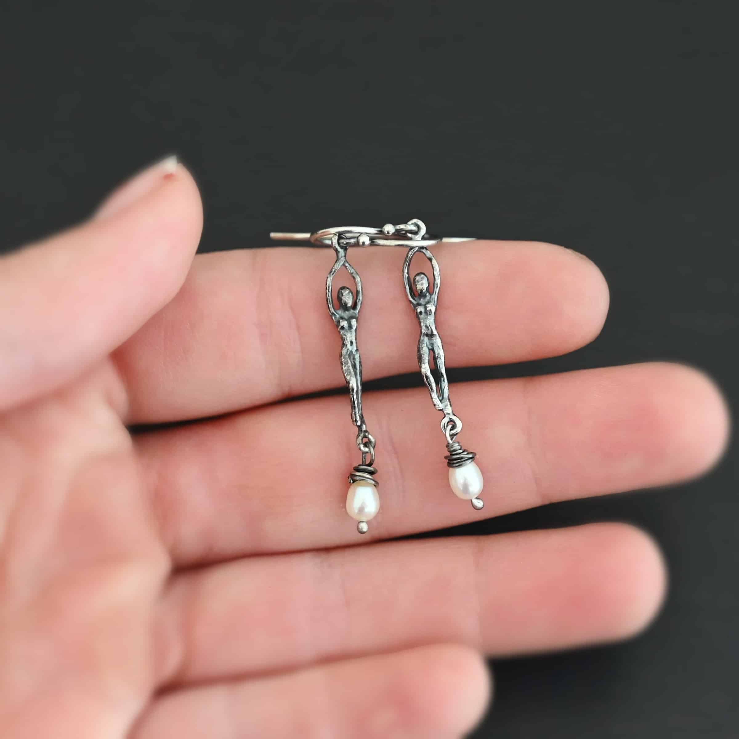 Close up of the "Goddess" Earrings. These earrings are made out of sterling silver and pearls with french hook ear wires. The earrings are sterling silver "women". The pearl hangs from the bottom of the earrings. The pearl is wrapped with sterling silver that is shiny. The rest of the sterling silver is oxidized. Earrings are shown on the palm of a hand to demonstrate size.