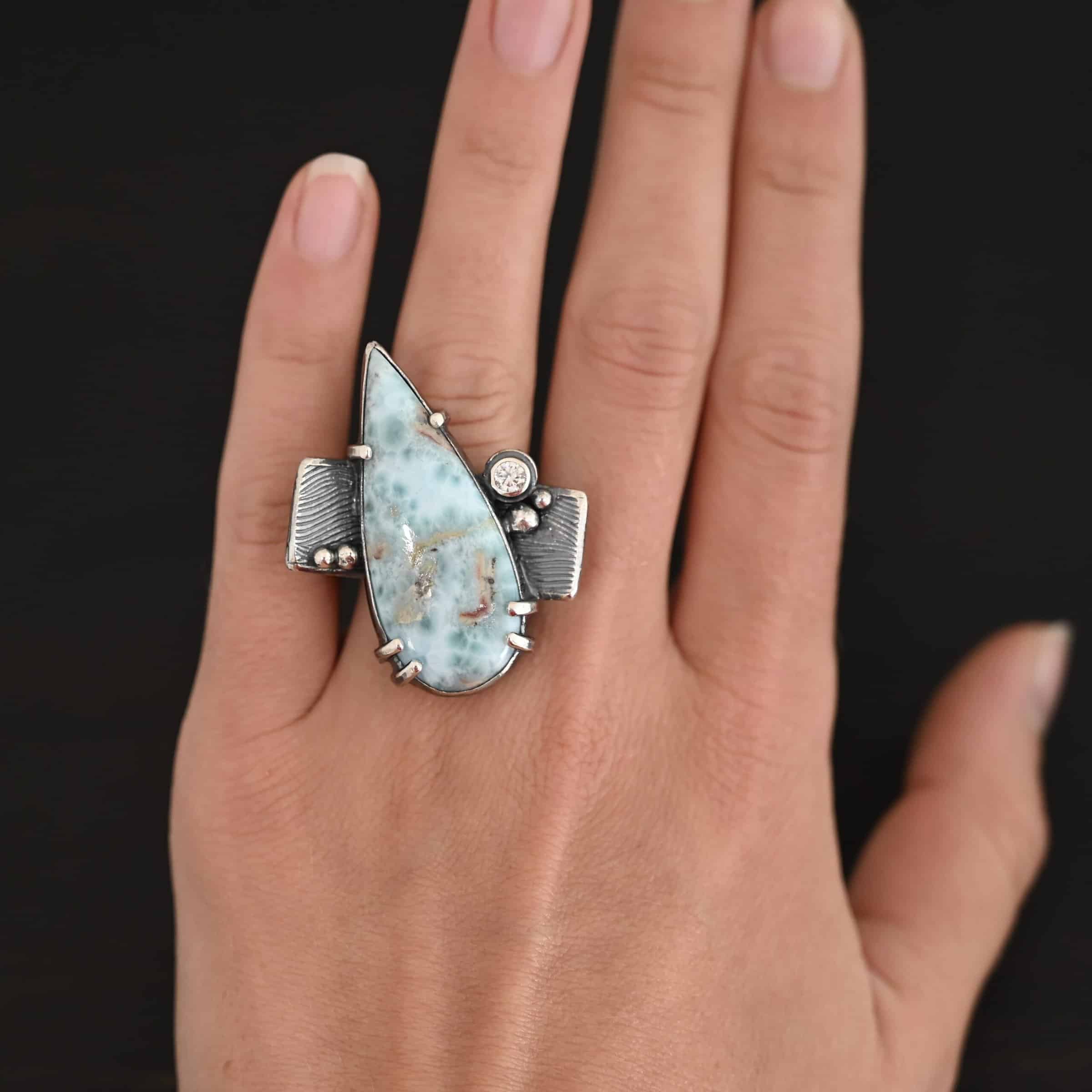 Just Add Water Ring Photo of the "Just Add Water" Ring. There is a large, light blue, teardrop shaped larimar in the center of the large, square ring shank. The stone is set sideways, so the point of the teardrop is pointing towards the top left. There is a moissanite on the top right side of the square ring shank. There are four balls of silver, two on the right side and two on the left side of the ring shank. The larimar has six silver prongs holding it in, four on the bottom and two on the top. Ring is worn by a model.