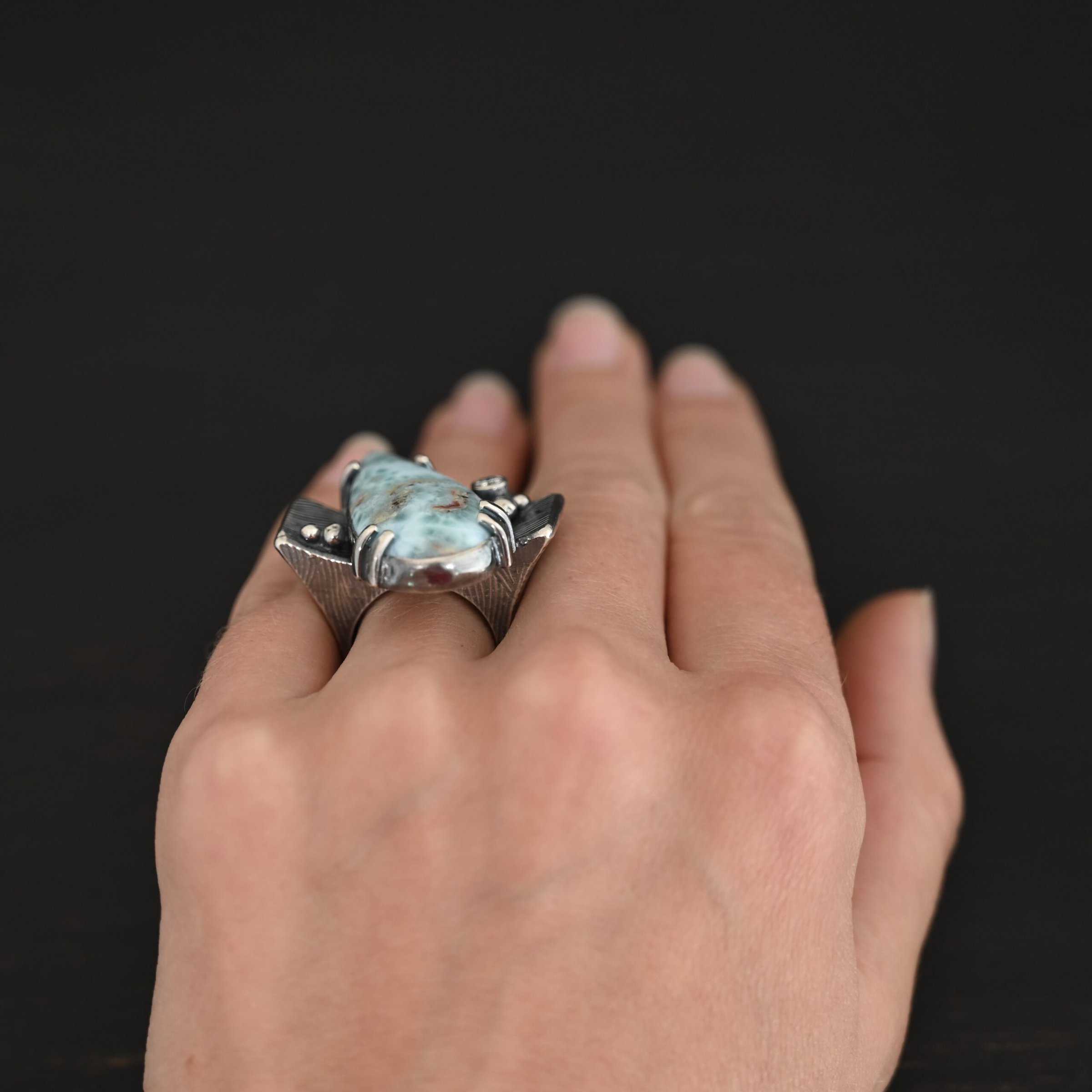 Just Add Water Ring Photo of the "Just Add Water" Ring. There is a large, light blue, teardrop shaped larimar in the center of the large, square ring shank. The stone is set sideways, so the point of the teardrop is pointing towards the top left. There is a moissanite on the top right side of the square ring shank. There are four balls of silver, two on the right side and two on the left side of the ring shank. The larimar has six silver prongs holding it in, four on the bottom and two on the top. Ring is worn by a model.