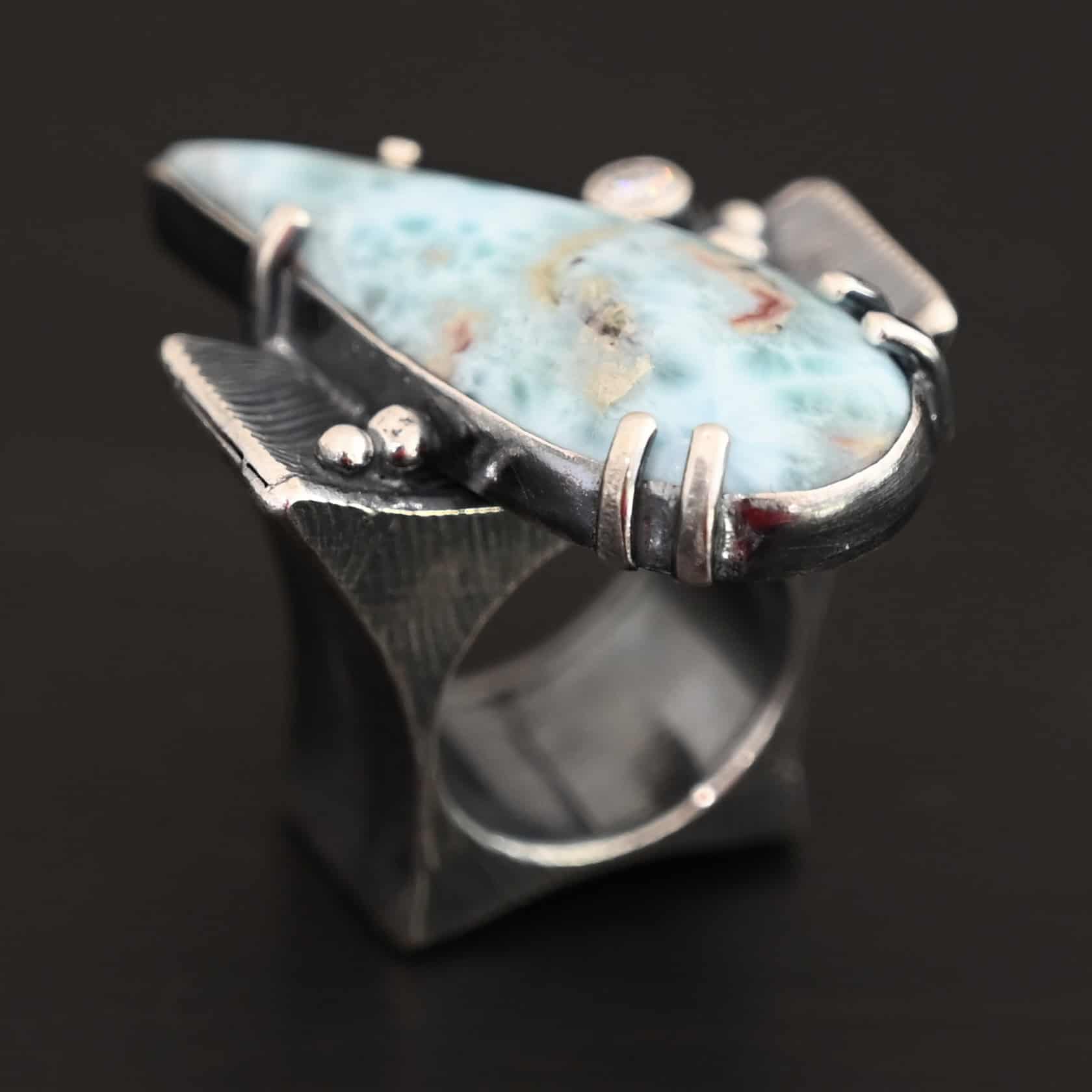 Just Add Water Ring Photo of the "Just Add Water" Ring. There is a large, light blue, teardrop shaped larimar in the center of the large, square ring shank. The stone is set sideways, so the point of the teardrop is pointing towards the top left. There is a moissanite on the top right side of the square ring shank. There are four balls of silver, two on the right side and two on the left side of the ring shank. The larimar has six silver prongs holding it in, four on the bottom and two on the top.