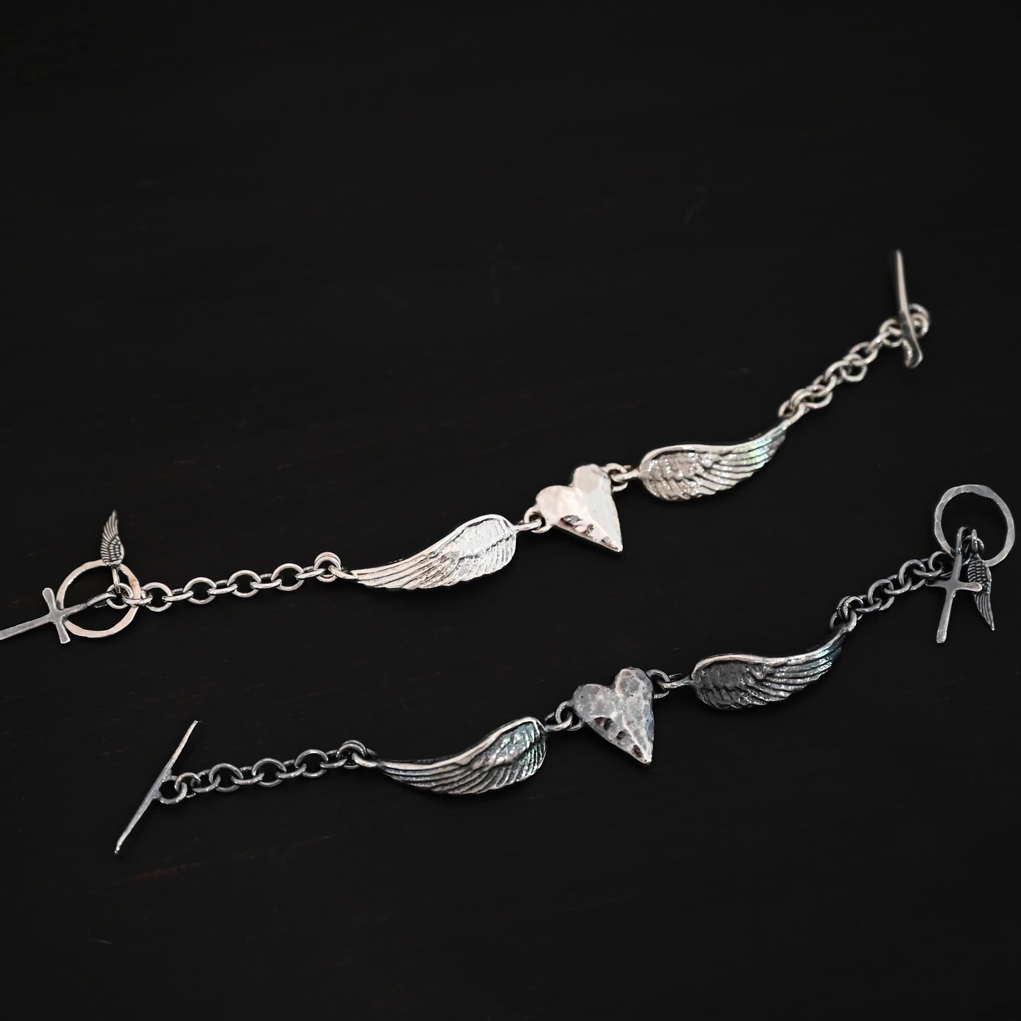Courage Bracelet "Courage" Bracelet as shown in shiny and oxidized. Bracelet is made out of solid sterling silver with a casted heart and two angel wings. Casted heart is solid sterling silver, and connected by an angel wing on each side. Angel wings are connected to a chain with a toggle clasp to connect the bracelet together. One bracelet is shiny and the other is oxidized. On both bracelets the heart is hammered to give it a textured look. Bracelets are set against a dark background.