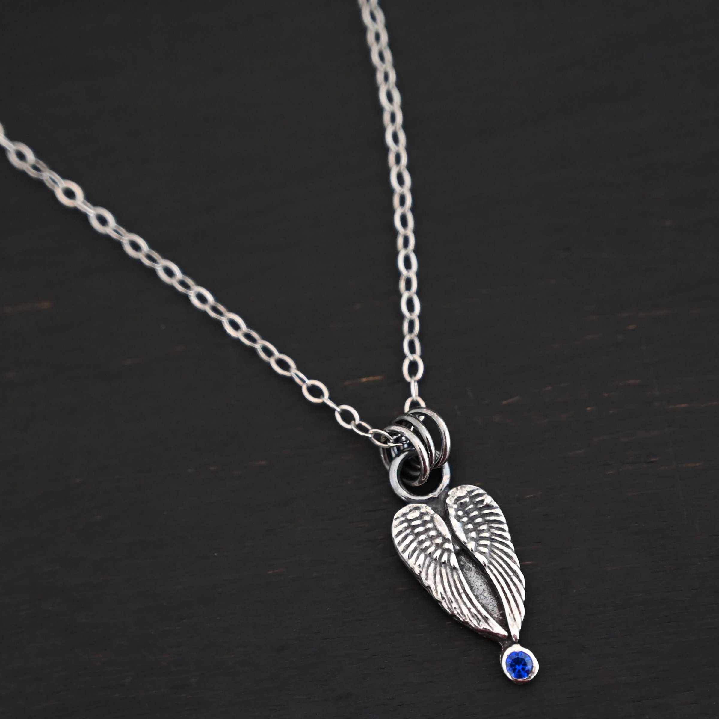 Close up of the "Strength" Birthstone Necklace. Shown as sterling silver, oxidized, and with an sapphire for the birthstone. Necklace is set against a dark background.