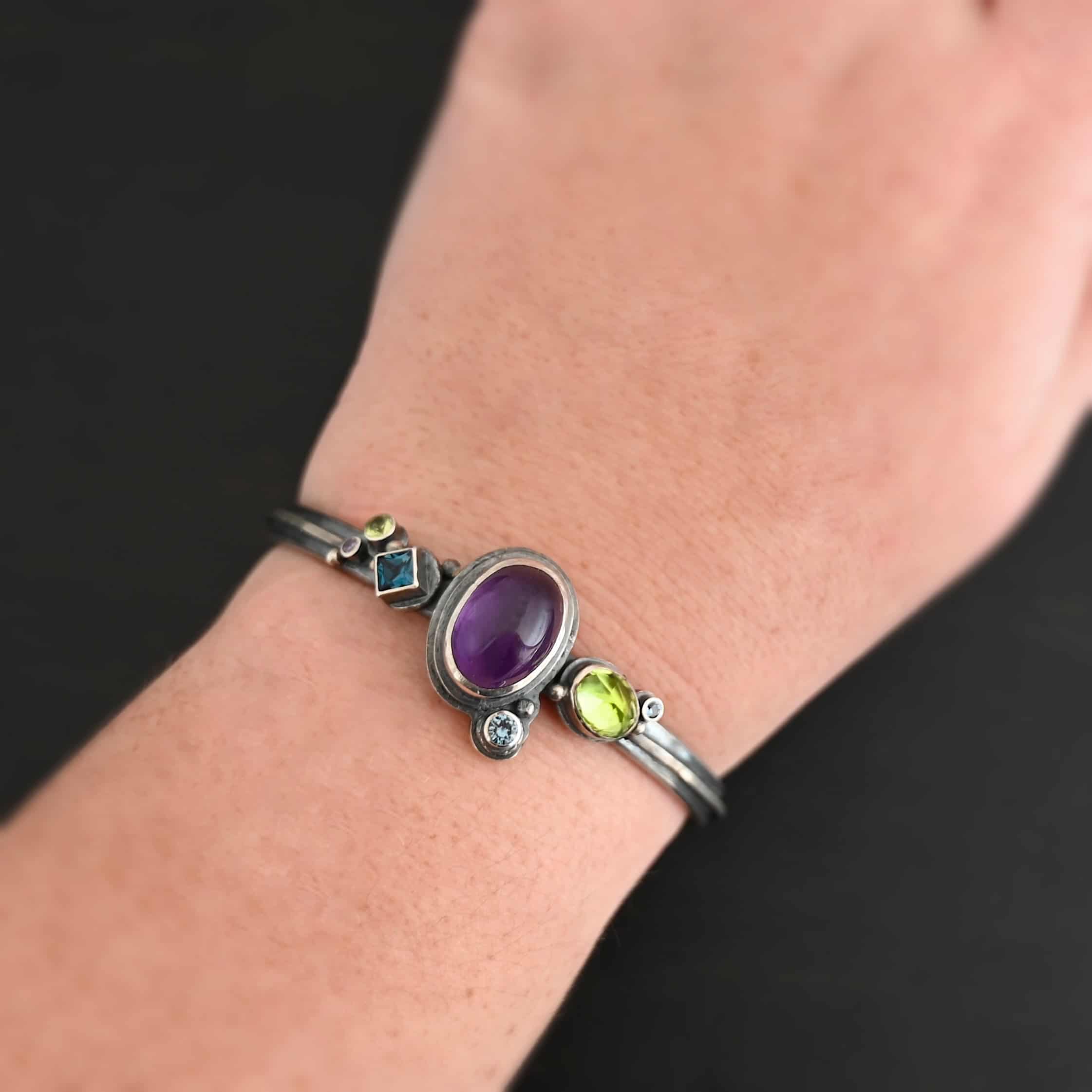 Springtime Violets Cuff "Springtime Violets" Cuff made out of Sterling Silver Amethyst, Peridot, Aquamarine, & Blue Zircon with the oval amethyst as the center stone and a large oval peridot next to it. There is a small circular aquamarine on the bottom close to the aquamarine. There is a square shaped Blue Zircon, a small circular peridot and small circular aquamarine on the left side of the bracelet. There is a model wearing the cuff on a wrist, and is resting against a dark background.