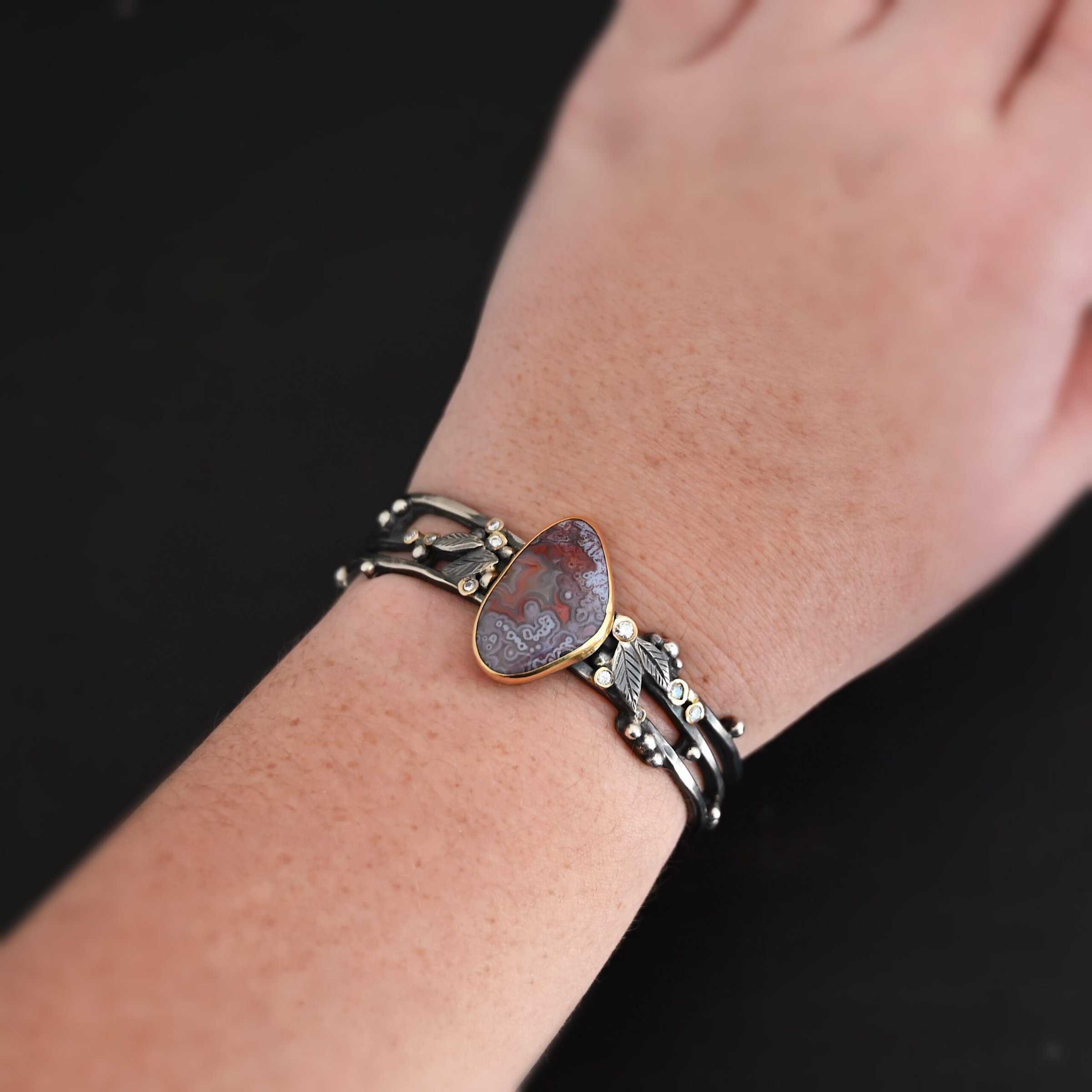 Aspen Cuff "Aspen" Cuff. This cuff is made out of sterling silver, gold, crazy lace agate, and moissanites. The oval shaped crazy lace agate is the center stone, with ten moissanites scattered around the cuff. There are four silver leaves on the cuff, two on each side of the agate. There are many small silver dots scattered around the cuff as well. The crazy lace agate and moissanites are all set in gold. The agate is a mix of red and purple swirls. The band is made out of three separate, but thick strands of sterling silver that are hammered. There is a model wearing a cuff on their wrist to show how large the cuff is. The hand and bracelet are set against a dark background.