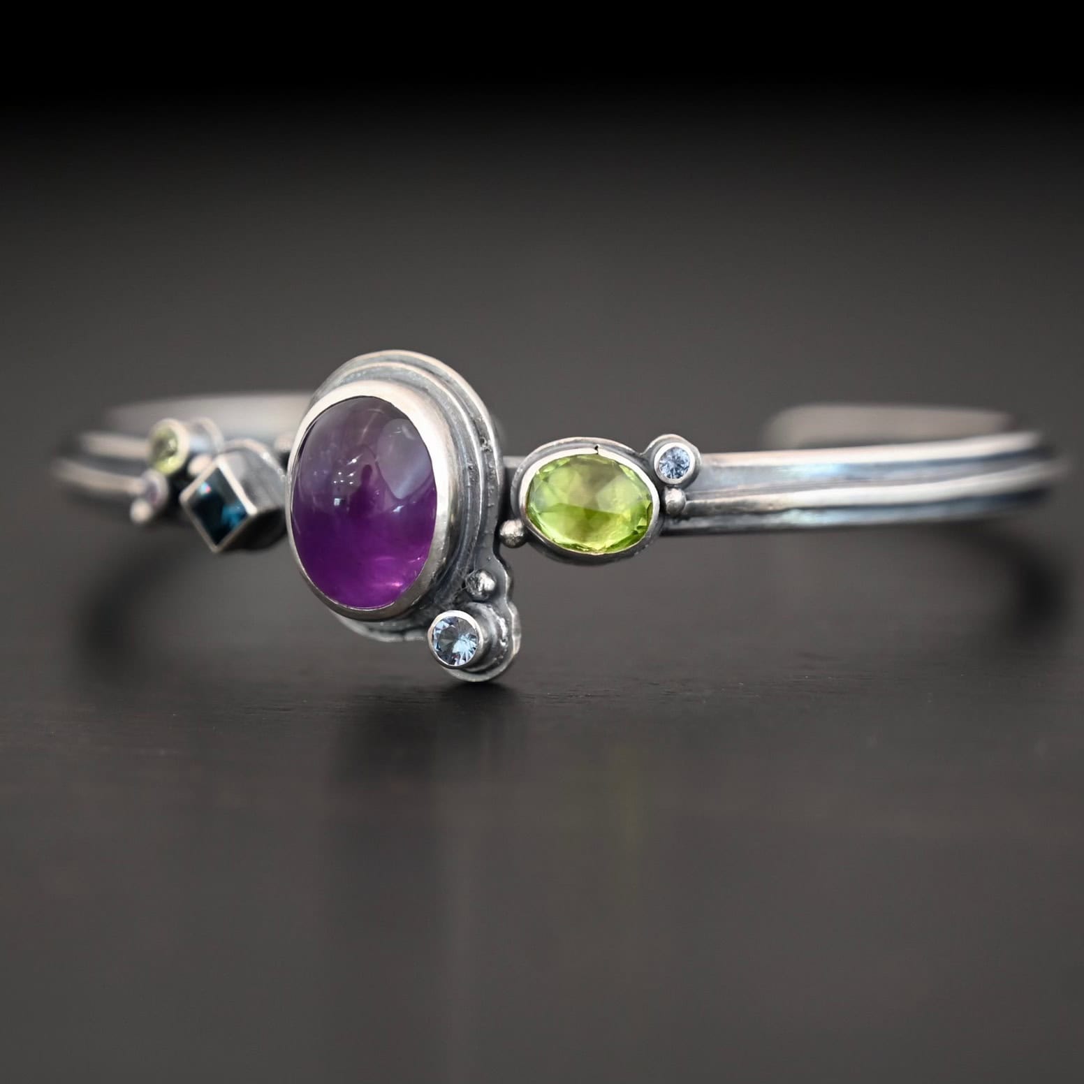 Springtime Violets Side view of the Springtime Violets Cuff made out of Sterling Silver Amethyst, Peridot, Aquamarine, & Blue Zircon with the oval amethyst as the center stone and a large oval peridot next to it. There is a small circular aquamarine on the bottom close to the aquamarine. There is a blue zircon on the left side of the cuff, along with another peridot and amethyst.