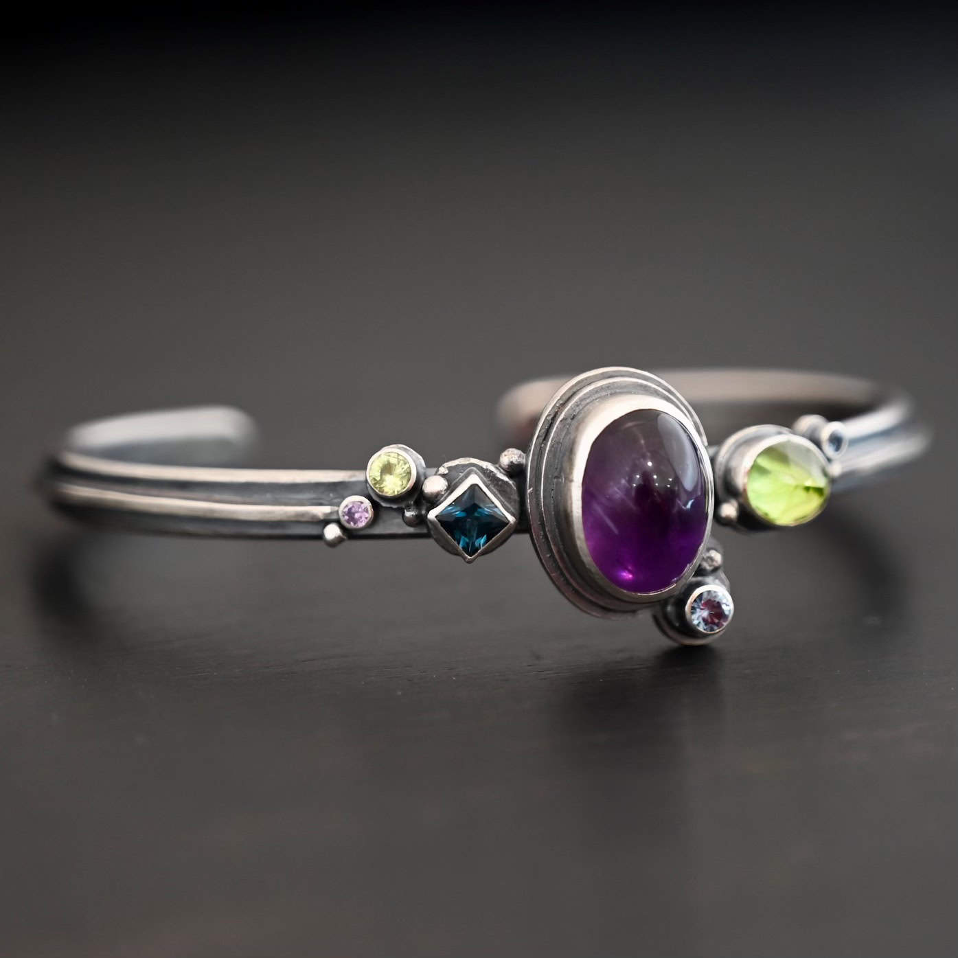 Springtime Violets Cuff Side view of the Springtime Violets Cuff made out of Sterling Silver Amethyst, Peridot, Aquamarine, & Blue Zircon with the oval amethyst as the center stone and a large oval peridot next to it. There is a small circular aquamarine on the bottom close to the aquamarine. The Blue Zircon, and another peridot and aquamarine are on the left side of the bracelet.