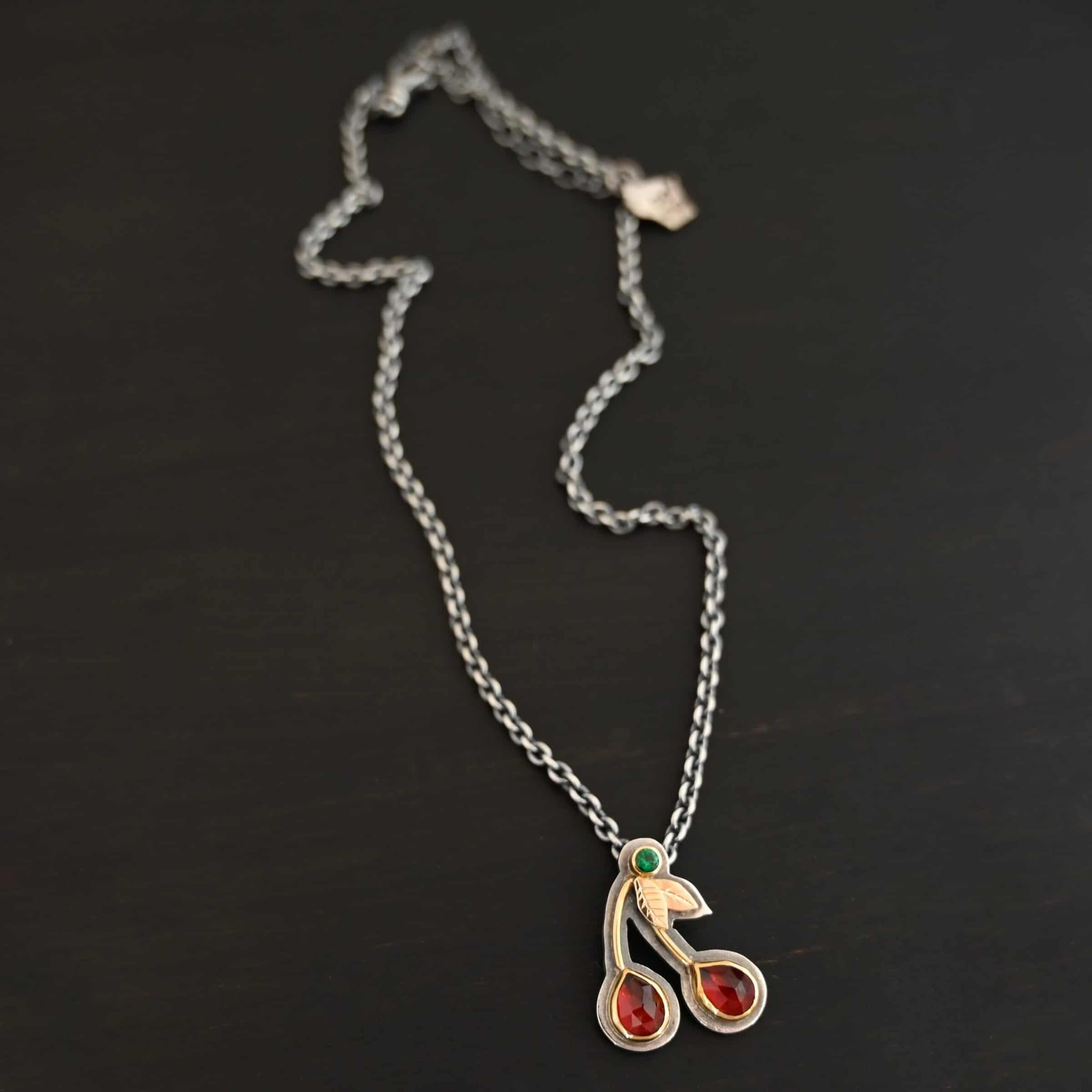Door County Cherry Necklace Door County Cherry Necklace made out of Sterling Silver, Gold, Rubies, & Tsavorite Garnet to look like cherries as the pendant of the necklace and it is on a 16-19" Savannah Chain. The necklace is laying against a dark background.