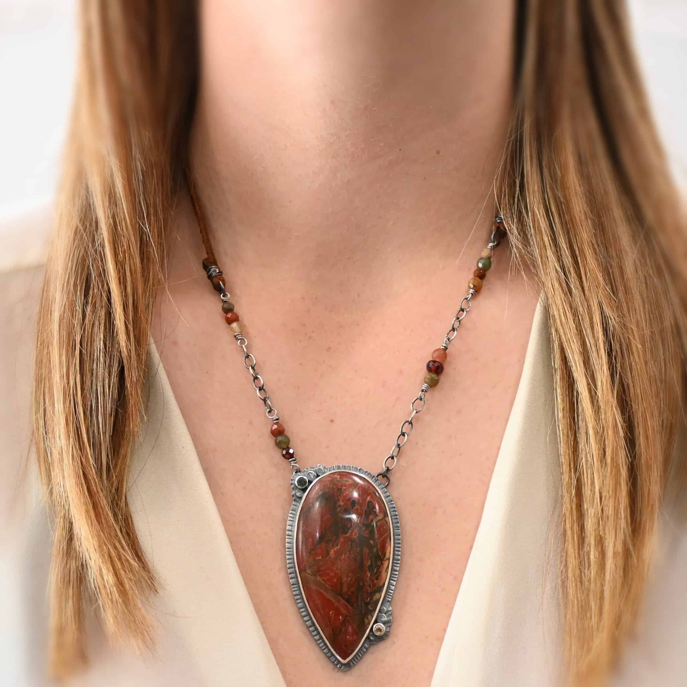 Sunset Orchard Necklace Close up of the "Sunset Orchard" Necklace. This necklace is made out of sterling silver, cherry creek jasper, and tourmaline. The cherry creek jasper is a large teardrop shape in a sterling silver setting, and there are two tourmalines, one on top of the pendant, and one closer to the bottom. The pendant is attached to a sterling silver chain with beads on both sides. There is a model wearing the necklace to demonstrate how big it is, and the model is wearing a white V-Neck shirt.