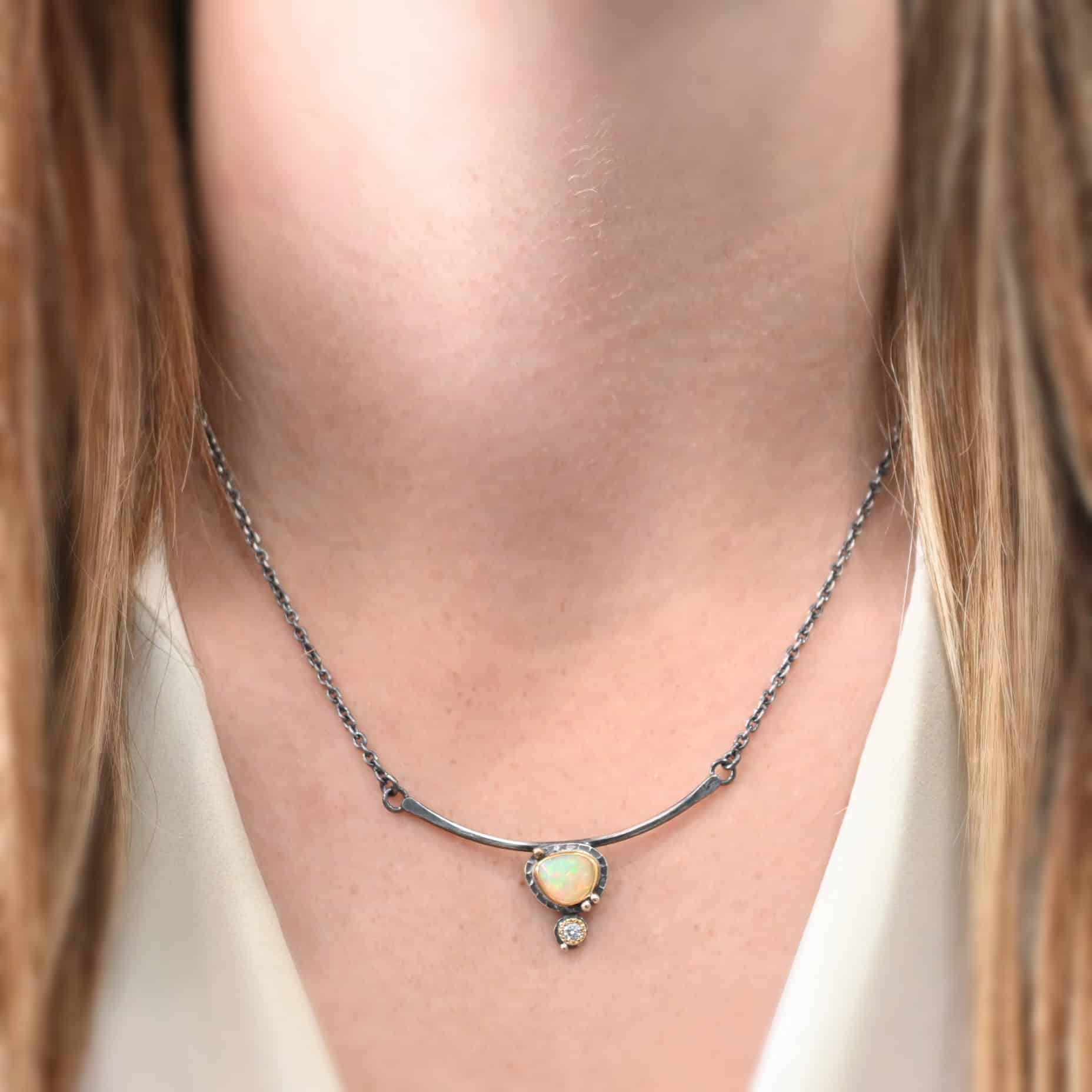The "Let The Sunshine Through Necklace" is made out of sterling silver, gold, opal, and moissanite. The opal is the center stone, with the moissanite attached directly below the opal. The opal and moissanite are attached to a curved bar, that goes two and a half inches across the neck. The necklace is on a model who is wearing a white V-Neck shirt.