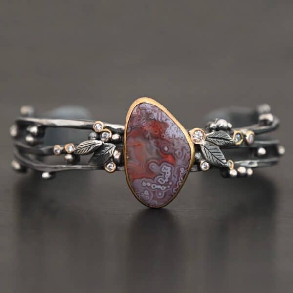 Close up of the "Aspen" Cuff. This cuff is made out of sterling silver, gold, crazy lace agate, and moissanites. The oval shaped crazy lace agate is the center stone, with ten moissanites scattered around the cuff. There are four silver leaves on the cuff, two on each side of the agate. There are many small silver dots scattered around the cuff as well. The crazy lace agate and moissanites are all set in gold. The agate is a mix of red and purple swirls. The band is made out of three separate, but thick strands of sterling silver that are hammered. The cuff is set against a dark background.