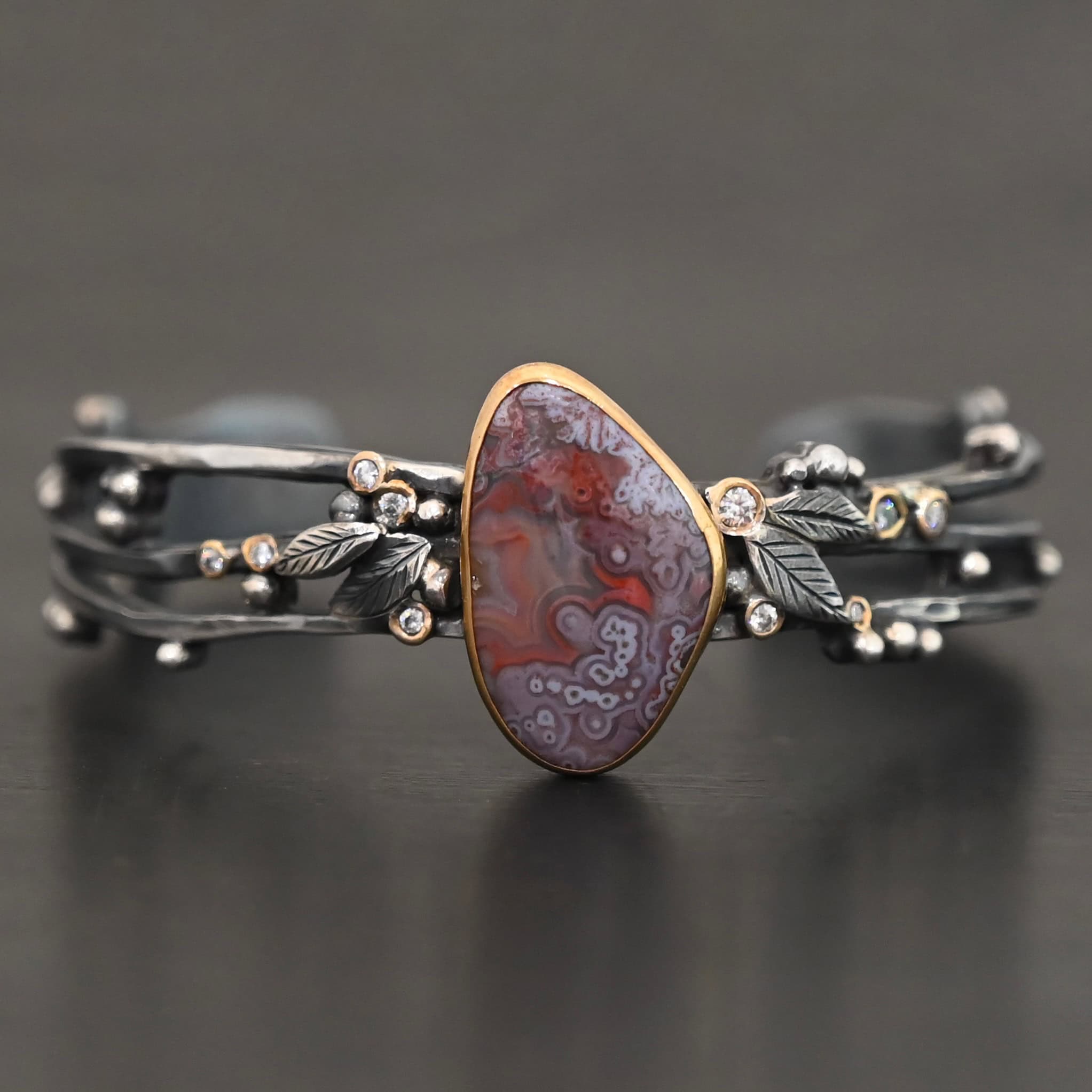 Aspen Cuff New Close up of the "Aspen" Cuff. This cuff is made out of sterling silver, gold, crazy lace agate, and moissanites. The oval shaped crazy lace agate is the center stone, with ten moissanites scattered around the cuff. There are four silver leaves on the cuff, two on each side of the agate. There are many small silver dots scattered around the cuff as well. The crazy lace agate and moissanites are all set in gold. The agate is a mix of red and purple swirls. The band is made out of three separate, but thick strands of sterling silver that are hammered. The cuff is set against a dark background.