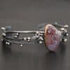 Close up of the "Aspen" Cuff. This cuff is made out of sterling silver, gold, crazy lace agate, and moissanites. The oval shaped crazy lace agate is the center stone, with ten moissanites scattered around the cuff. There are four silver leaves on the cuff, two on each side of the agate. There are many small silver dots scattered around the cuff as well. The crazy lace agate and moissanites are all set in gold. The agate is a mix of red and purple swirls. The band is made out of three separate, but thick strands of sterling silver that are hammered. The cuff is set against a dark background.