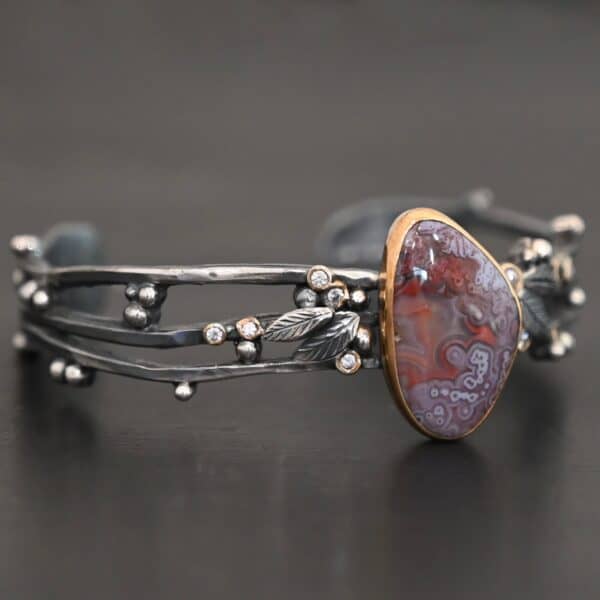 Close up of the "Aspen" Cuff. This cuff is made out of sterling silver, gold, crazy lace agate, and moissanites. The oval shaped crazy lace agate is the center stone, with ten moissanites scattered around the cuff. There are four silver leaves on the cuff, two on each side of the agate. There are many small silver dots scattered around the cuff as well. The crazy lace agate and moissanites are all set in gold. The agate is a mix of red and purple swirls. The band is made out of three separate, but thick strands of sterling silver that are hammered. The cuff is set against a dark background.