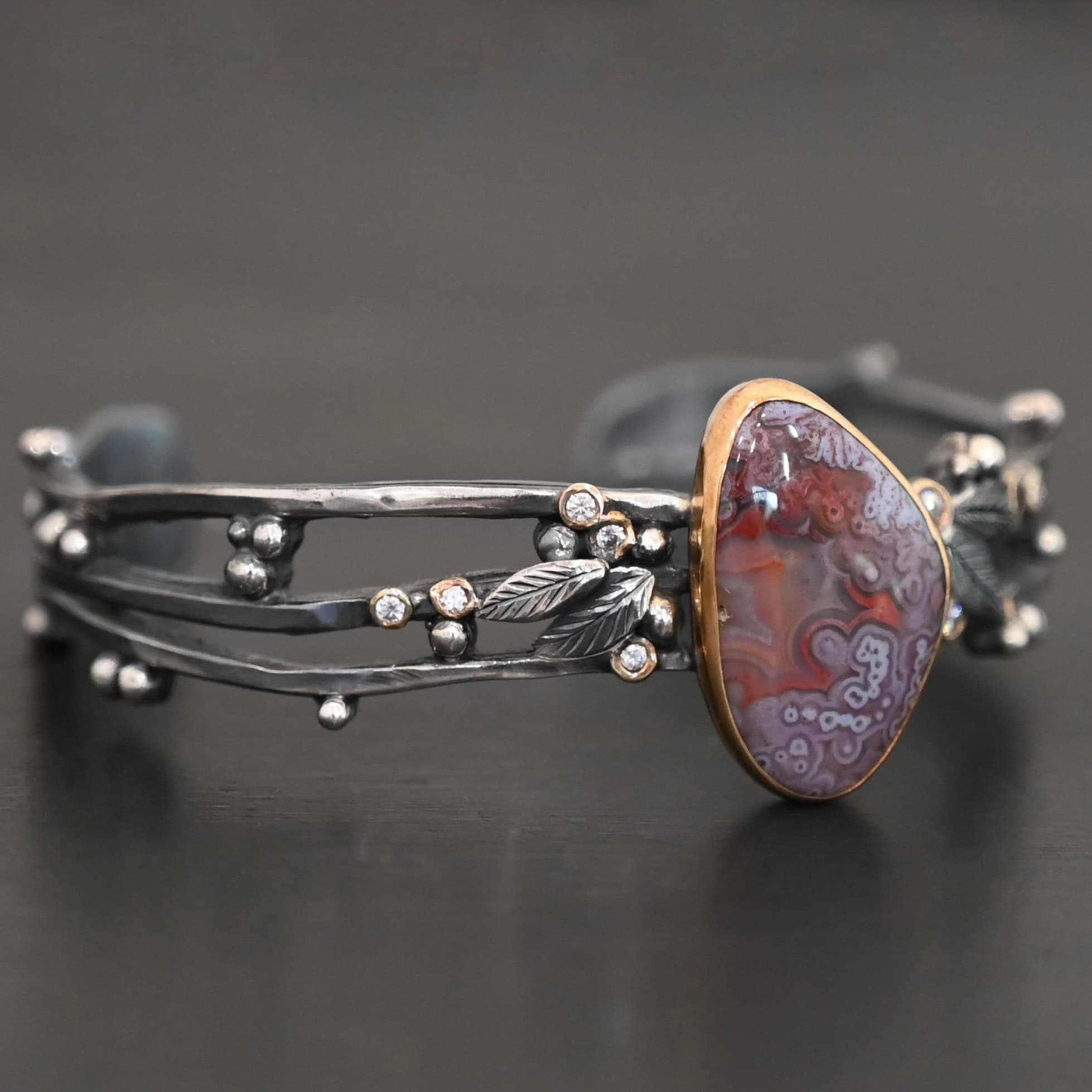 Aspen Cuff New Close up of the "Aspen" Cuff. This cuff is made out of sterling silver, gold, crazy lace agate, and moissanites. The oval shaped crazy lace agate is the center stone, with ten moissanites scattered around the cuff. There are four silver leaves on the cuff, two on each side of the agate. There are many small silver dots scattered around the cuff as well. The crazy lace agate and moissanites are all set in gold. The agate is a mix of red and purple swirls. The band is made out of three separate, but thick strands of sterling silver that are hammered. The cuff is set against a dark background.