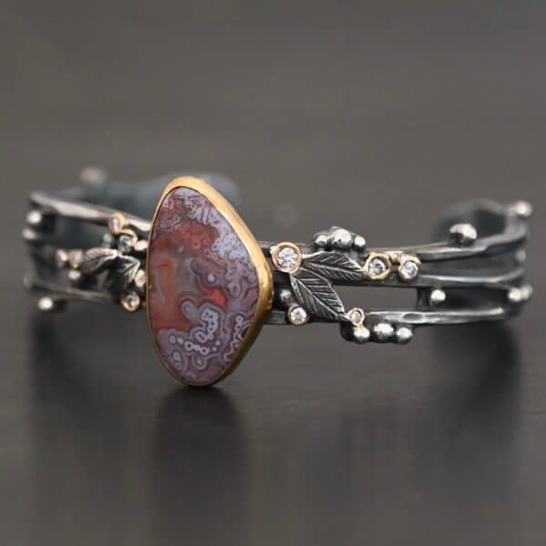 Close up of the "Aspen" Cuff. This cuff is made out of sterling silver, gold, crazy lace agate, and moissanites. The oval shaped crazy lace agate is the center stone, with ten moissanites scattered around the cuff. There are four silver leaves on the cuff, two on each side of the agate. There are many small silver dots scattered around the cuff as well. The crazy lace agate and moissanites are all set in gold. The agate is a mix of red and purple swirls. The band is made out of three separate, but thick strands of sterling silver that are hammered. The cuff is set against a dark background.