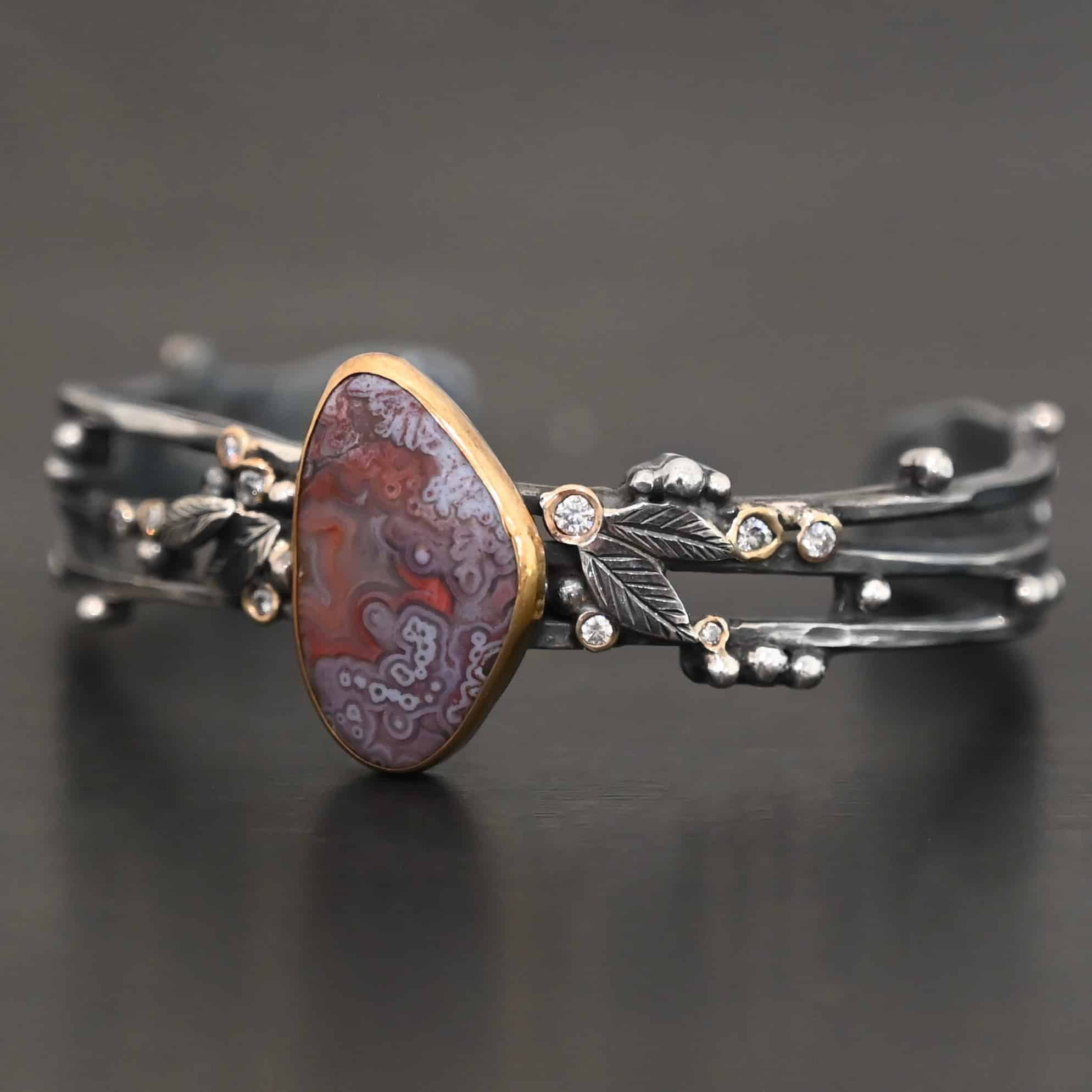 Aspen Cuff New Close up of the "Aspen" Cuff. This cuff is made out of sterling silver, gold, crazy lace agate, and moissanites. The oval shaped crazy lace agate is the center stone, with ten moissanites scattered around the cuff. There are four silver leaves on the cuff, two on each side of the agate. There are many small silver dots scattered around the cuff as well. The crazy lace agate and moissanites are all set in gold. The agate is a mix of red and purple swirls. The band is made out of three separate, but thick strands of sterling silver that are hammered. The cuff is set against a dark background.