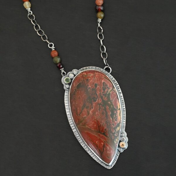 "Sunset Orchard" Necklace. This necklace is made out of sterling silver, cherry creek jasper, and tourmaline. The cherry creek jasper is a large teardrop shape in a sterling silver setting, and there are two tourmalines, one on top of the pendant, and one closer to the bottom. The pendant is attached to a sterling silver chain with beads on both sides.