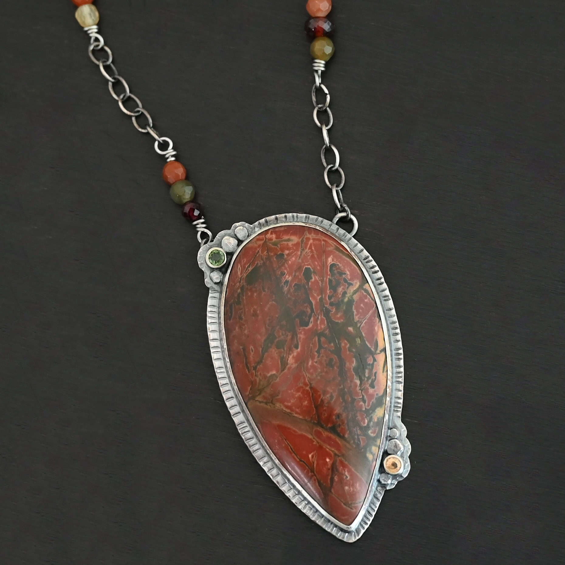 Sunset Orchard Necklace "Sunset Orchard" Necklace. This necklace is made out of sterling silver, cherry creek jasper, and tourmaline. The cherry creek jasper is a large teardrop shape in a sterling silver setting, and there are two tourmalines, one on top of the pendant, and one closer to the bottom. The pendant is attached to a sterling silver chain with beads on both sides.