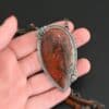 Close up of the "Sunset Orchard" Necklace. This necklace is made out of sterling silver, cherry creek jasper, and tourmaline. The cherry creek jasper is a large teardrop shape in a sterling silver setting, and there are two tourmalines, one on top of the pendant, and one closer to the bottom. The pendant is attached to a sterling silver chain with beads on both sides. The pendant is laying in the fingertips of a hand that is resting against a dark background.