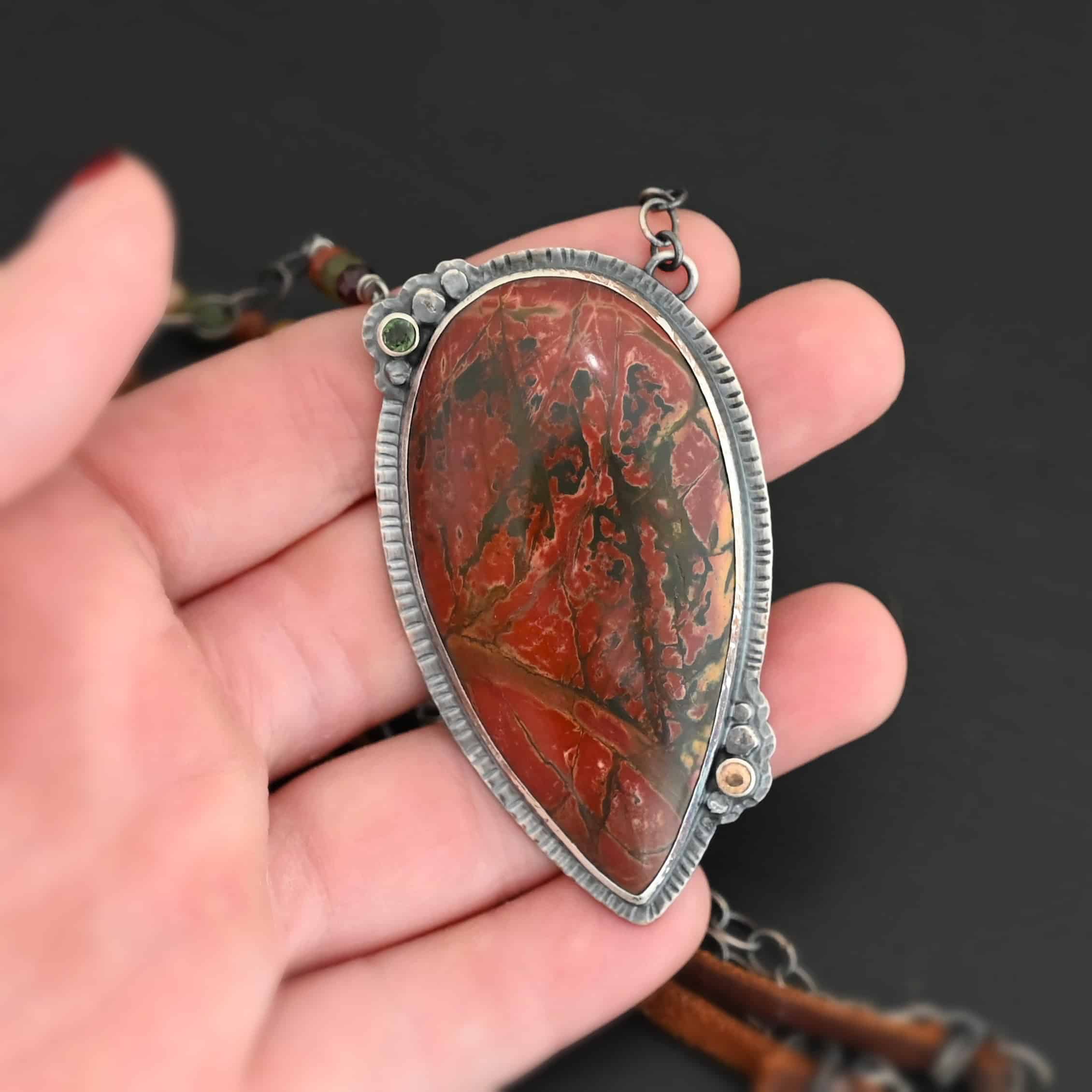 Sunset Orchard Necklace Close up of the "Sunset Orchard" Necklace. This necklace is made out of sterling silver, cherry creek jasper, and tourmaline. The cherry creek jasper is a large teardrop shape in a sterling silver setting, and there are two tourmalines, one on top of the pendant, and one closer to the bottom. The pendant is attached to a sterling silver chain with beads on both sides. The pendant is laying in the fingertips of a hand that is resting against a dark background.
