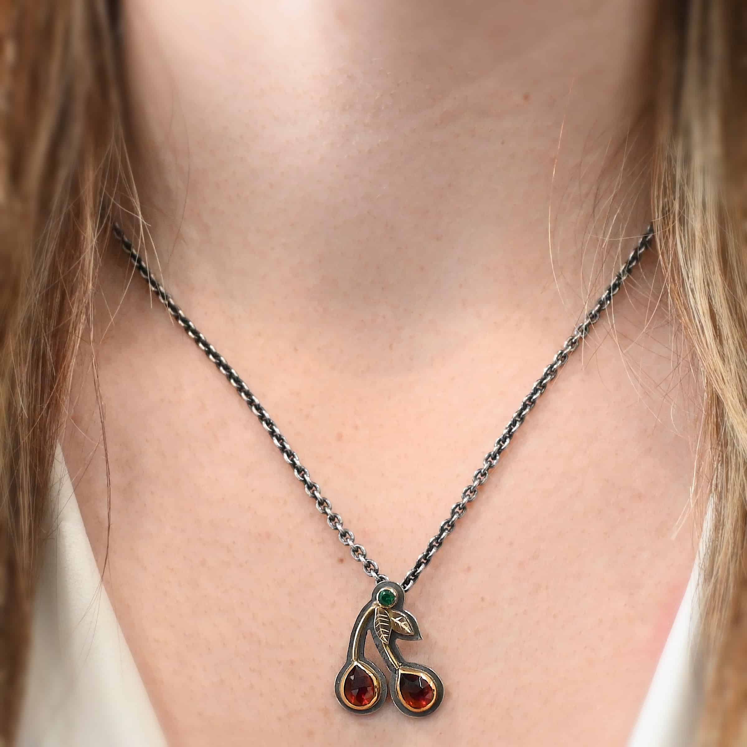 Door County Cherry Necklace Close up of the "Door County Cherry" Necklace. Made out of sterling silver, gold, garnets, and tsavorite garnet. There are two rubies that are both teardrop shaped with the points facing upwards. There are two small gold stems that lead to three small gold leaves and the tsavorite garnet that is set in gold. All of the stones are set in gold but have oxidized sterling silver around the gold. There is a loop on the back of the pendant that is attached to jump rings that are hanging on a chain. Necklace is shown on a model at 17" long to demonstrate length.