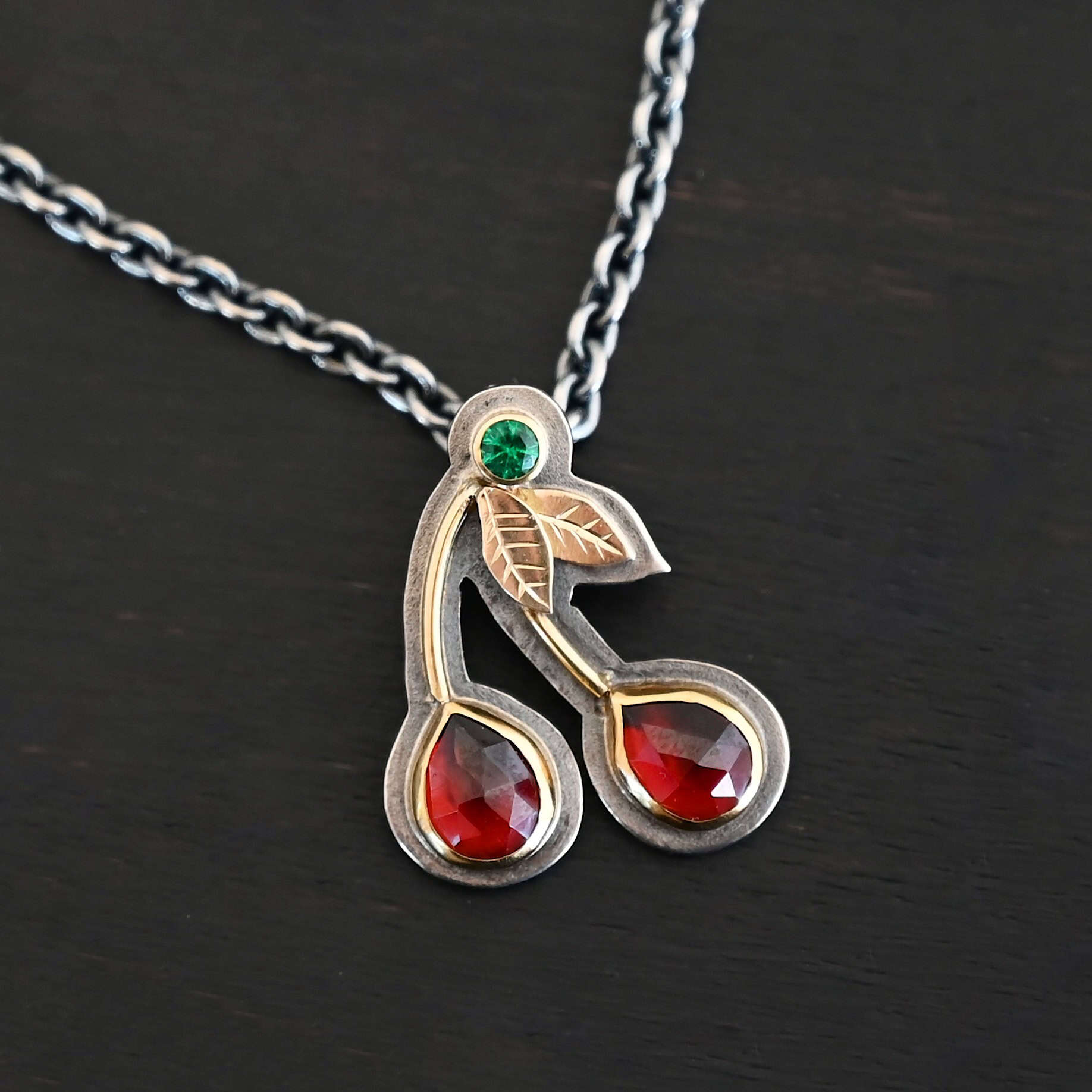 Door County Cherry Necklace New Close up of the "Door County Cherry" Necklace. Made out of sterling silver, gold, garnets, and tsavorite garnet. There are two rubies that are both teardrop shaped with the points facing upwards. There are two small gold stems that lead to three small gold leaves and the tsavorite garnet that is set in gold. All of the stones are set in gold but have oxidized sterling silver around the gold. There is a loop on the back of the pendant that is attached to jump rings that are hanging on a chain.