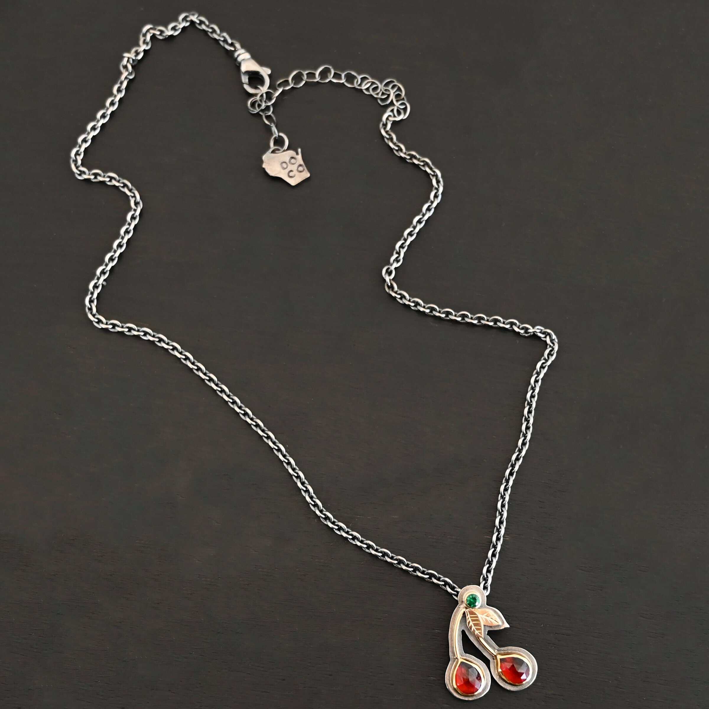Door County Cherry Necklace New Close up of the "Door County Cherry" Necklace. Made out of sterling silver, gold, garnets, and tsavorite garnet. There are two rubies that are both teardrop shaped with the points facing upwards. There are two small gold stems that lead to three small gold leaves and the tsavorite garnet that is set in gold. All of the stones are set in gold but have oxidized sterling silver around the gold. There is a loop on the back of the pendant that is attached to jump rings that are hanging on a chain.
