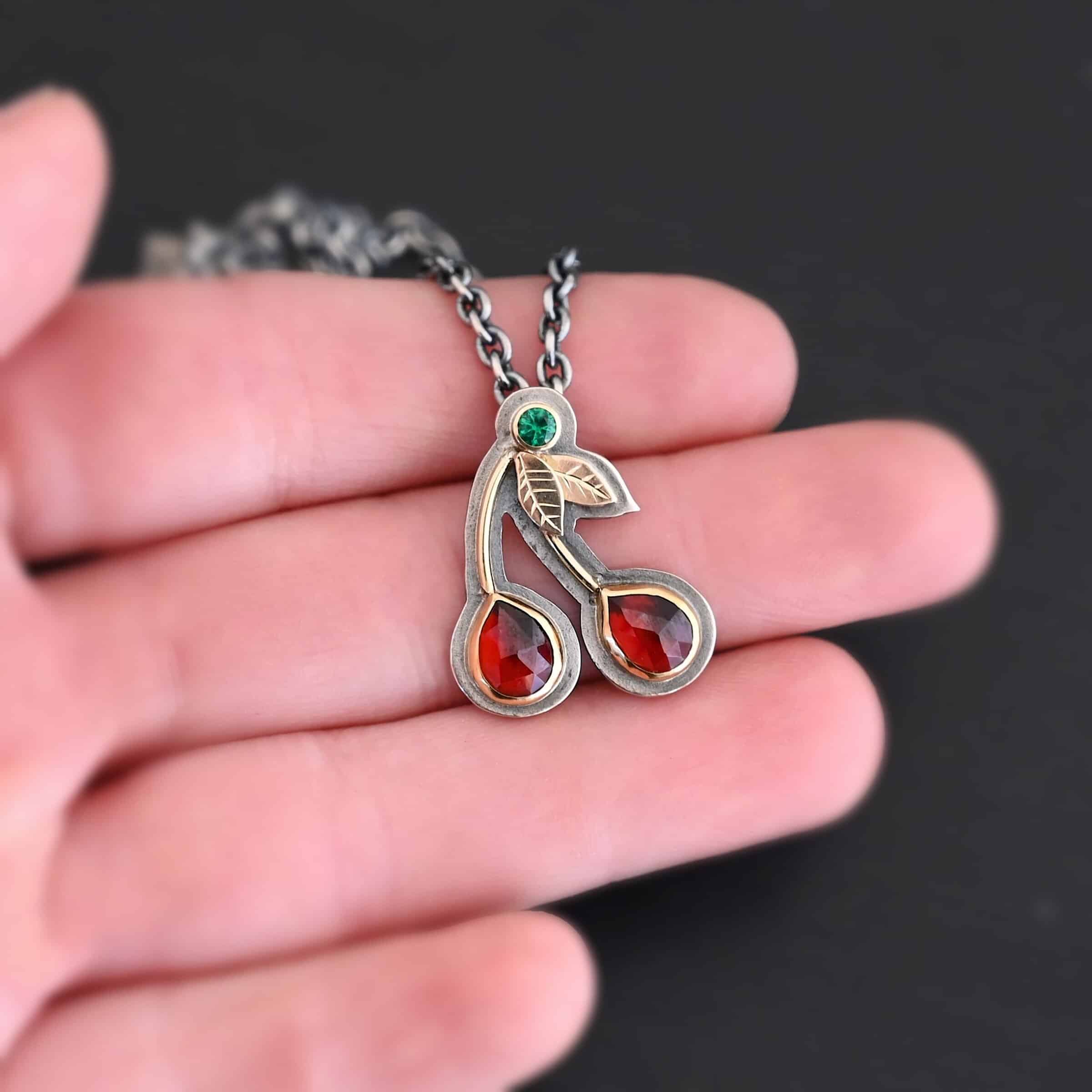 Door County Cherry Necklace New Door County Cherry Necklace made out of Sterling Silver, Gold, Rubies, & Tsavorite Garnet to look like cherries as the pendant of the necklace. The cherry pendant is laying on the fingers of a hand which is resting against a dark background