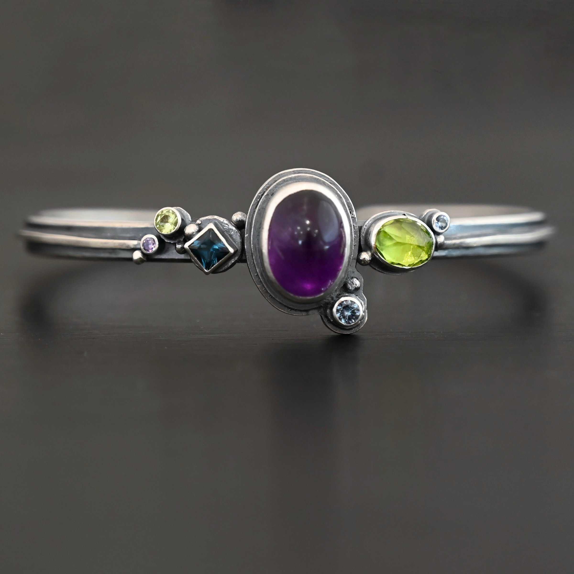 Springtime Violets Bracelet Close up of the "Springtime Violets" Cuff made out of Sterling Silver Amethyst, Peridot, Aquamarine, & Blue Zircon with the oval amethyst as the center stone and a large oval peridot next to it. There is a small circular aquamarine on the bottom close to the aquamarine. There is a square shaped Blue Zircon, a small circular peridot and small circular aquamarine on the left side of the bracelet. The cuff is set against a dark background.