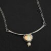 Close up of the "Let The Sunshine Through" Necklace This necklace is made out of sterling silver, gold, opal, and moissanite. The opal is the center stone, with the moissanite attached directly below the opal. There are four gold dots on the necklace, and they are scattered around the center opal with one on the bottom moissanite. The opal and moissanite are attached to a curved bar. The bar is attached on both sides to a Savannah chain to complete the necklace. The Savannah Chain is made out of small, but thick oval links. The necklace is set against a dark background.