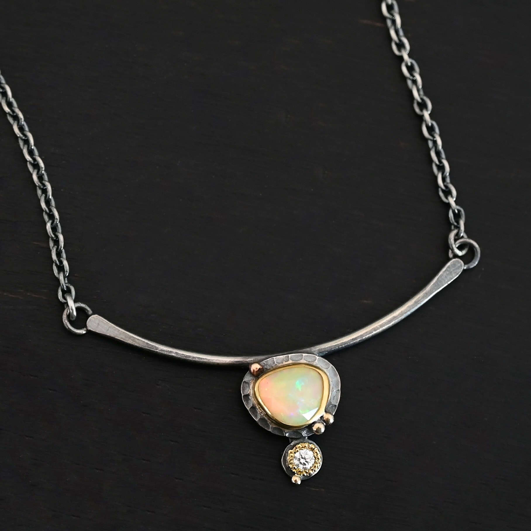 Close up of the "Let The Sunshine Through" Necklace This necklace is made out of sterling silver, gold, opal, and moissanite. The opal is the center stone, with the moissanite attached directly below the opal. There are four gold dots on the necklace, and they are scattered around the center opal with one on the bottom moissanite. The opal and moissanite are attached to a curved bar. The bar is attached on both sides to a Savannah chain to complete the necklace. The Savannah Chain is made out of small, but thick oval links. The necklace is set against a dark background.