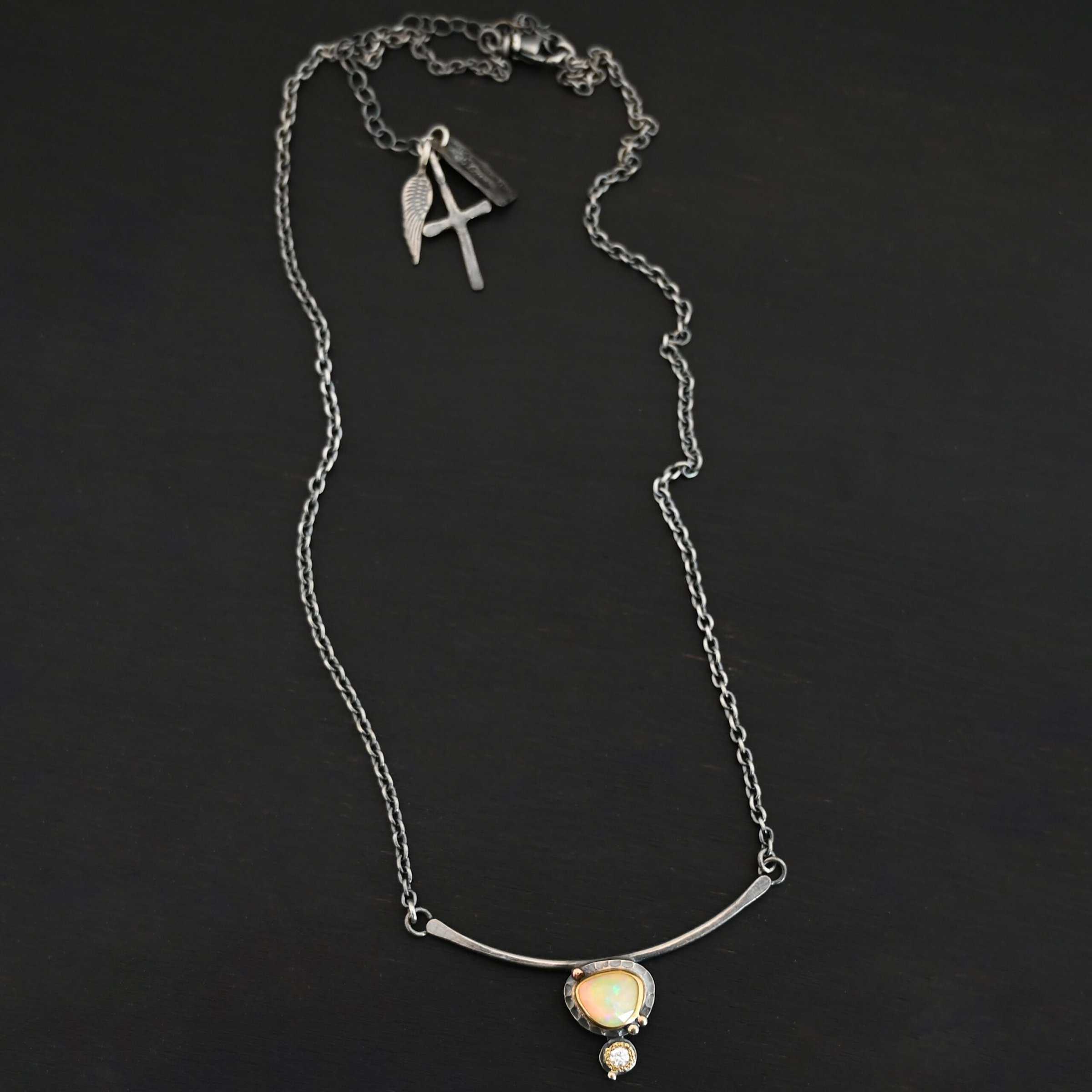 Close up of the "Let The Sunshine Through" Necklace This necklace is made out of sterling silver, gold, opal, and moissanite. The opal is the center stone, with the moissanite attached directly below the opal. There are four gold dots on the necklace, and they are scattered around the center opal with one on the bottom moissanite. The opal and moissanite are attached to a curved bar. The bar is attached on both sides to a Savannah chain to complete the necklace. The Savannah Chain is made out of small, but thick oval links. The necklace is set against a dark background.