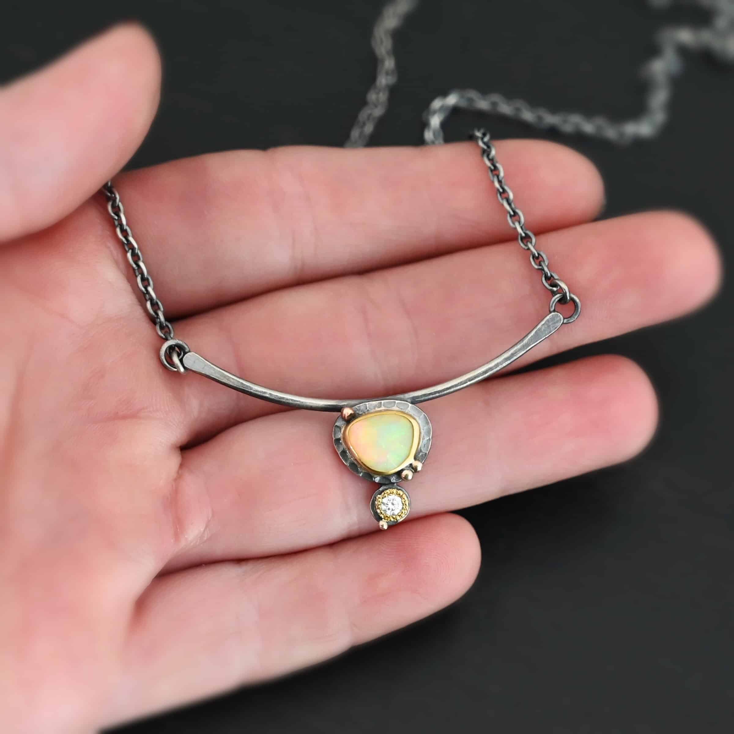 Close up of the "Let The Sunshine Through" Necklace This necklace is made out of sterling silver, gold, opal, and moissanite. The opal is the center stone, with the moissanite attached directly below the opal. There are four gold dots on the necklace, and they are scattered around the center opal with one on the bottom moissanite. The opal and moissanite are attached to a curved bar. The bar is attached on both sides to a Savannah chain to complete the necklace. The Savannah Chain is made out of small, but thick oval links. The necklace is set against the fingertips of a hand.