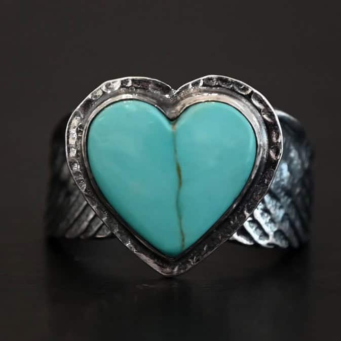 Untamable Spirit Ring Close up of the "Untamable Spirit" Ring. Made out of sterling silver and a heart shaped turquoise stone that is a teal color. It has a dark green but very thin stripe going down the center of the stone. The stone is set in sterling silver that is hammered around the edges. The band of the ring has two angel wings, one on each side that are curved and that touch at the back, but are not soldered together. Ring is set against a dark background.