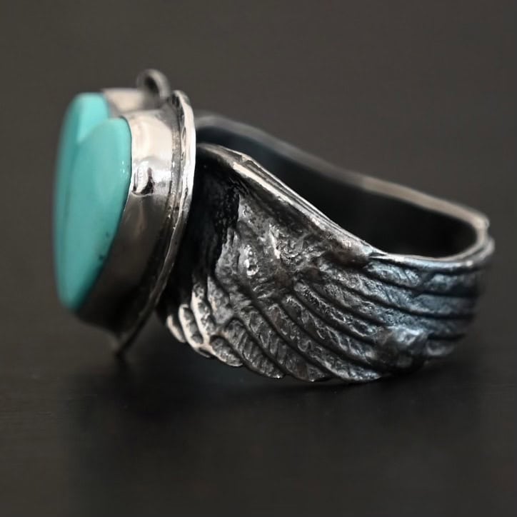 Untamable Spirit Ring Close up of the side of the "Untamable Spirit" Ring. The band is made out of a sterling silver angel wing that curves to go towards the back of the ring and meet the other side of the band. Ring is set against a dark background.