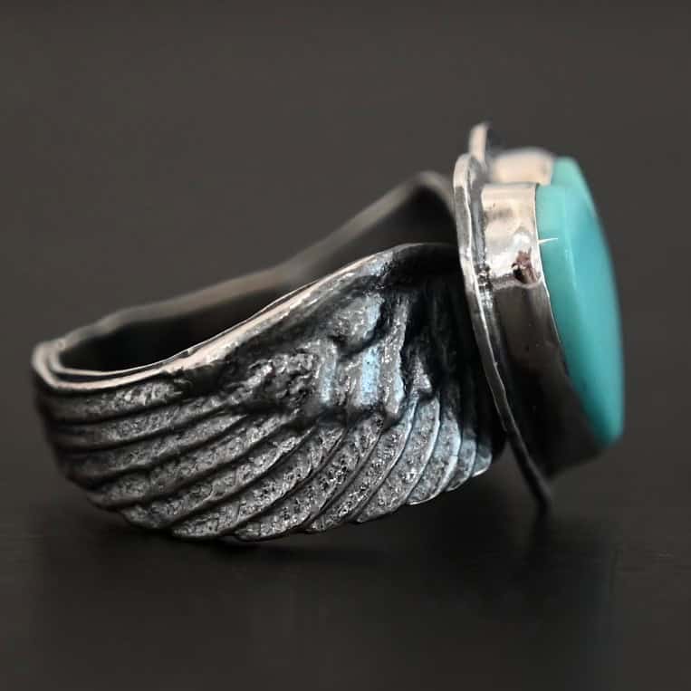 Untamable Spirit Ring Close up of the side of the "Untamable Spirit" Ring. The band is made out of a sterling silver angel wing that curves to go towards the back of the ring and meet the other side of the band. Ring is set against a dark background.