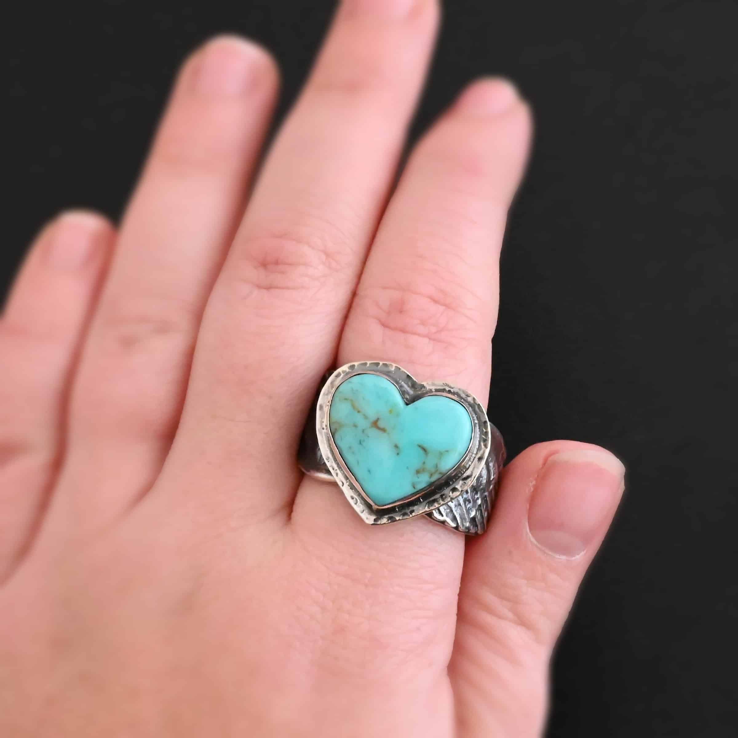 Untamable Spirit Ring New Close up of the "Untamable Spirit" Ring. Made out of sterling silver and a heart shaped turquoise stone that is a teal color. It has a dark green but very thin stripe going down the center of the stone. The stone is set in sterling silver that is hammered around the edges. The band of the ring has two angel wings, one on each side that are curved and that touch at the back, but are not soldered together. Ring is worn on a models pointer finger to demonstrate size.