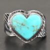 Close up of the "Untamable Spirit" Ring. Made out of sterling silver and a heart shaped turquoise stone that is a teal color. It has dark green but very thin stripes going around the stone. The stone is set in sterling silver that is hammered around the edges. The band of the ring has two angel wings, one on each side that are curved and that touch at the back, but are not soldered together. Ring is set against a dark background.