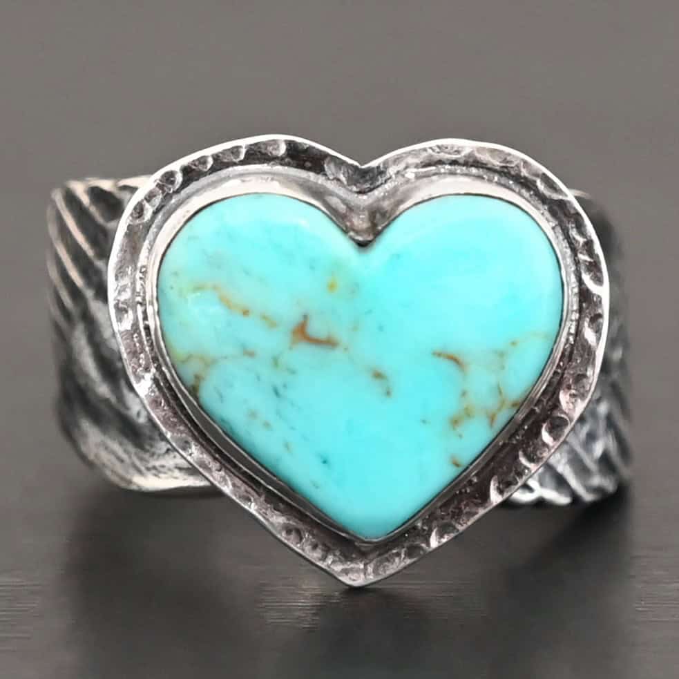 Untamable Spirit Ring Close up of the "Untamable Spirit" Ring. Made out of sterling silver and a heart shaped turquoise stone that is a teal color. It has dark green but very thin stripes going around the stone. The stone is set in sterling silver that is hammered around the edges. The band of the ring has two angel wings, one on each side that are curved and that touch at the back, but are not soldered together. Ring is set against a dark background.