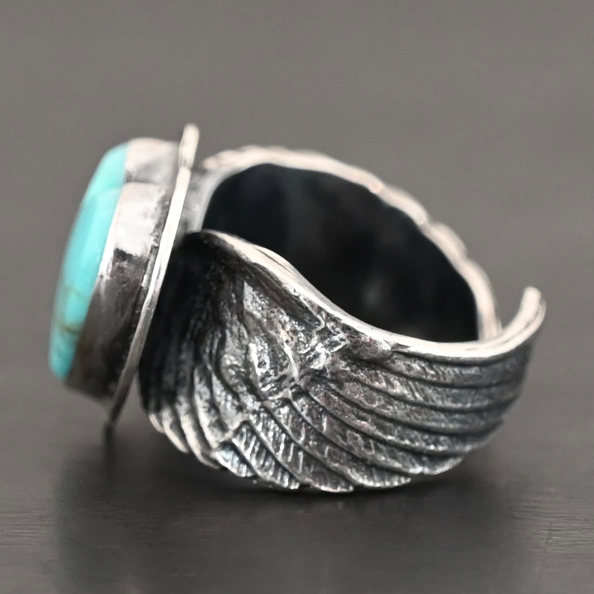 Untamable Spirit Ring Close up of the side of the "Untamable Spirit" Ring. The band is made out of a sterling silver angel wing that curves to go towards the back of the ring and meet the other side of the band. Ring is set against a dark background.