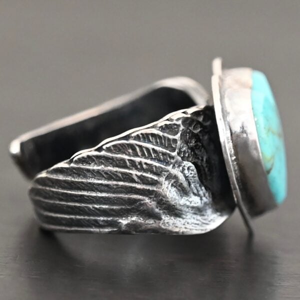 Close up of the side of the "Untamable Spirit" Ring. The band is made out of a sterling silver angel wing that curves to go towards the back of the ring and meet the other side of the band. Ring is set against a dark background.