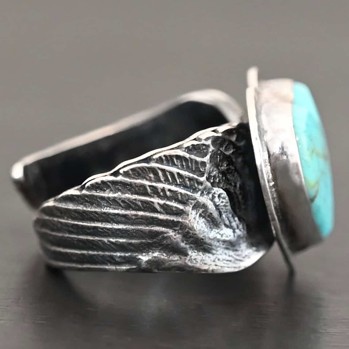 Untamable Spirit Ring Close up of the side of the "Untamable Spirit" Ring. The band is made out of a sterling silver angel wing that curves to go towards the back of the ring and meet the other side of the band. Ring is set against a dark background.