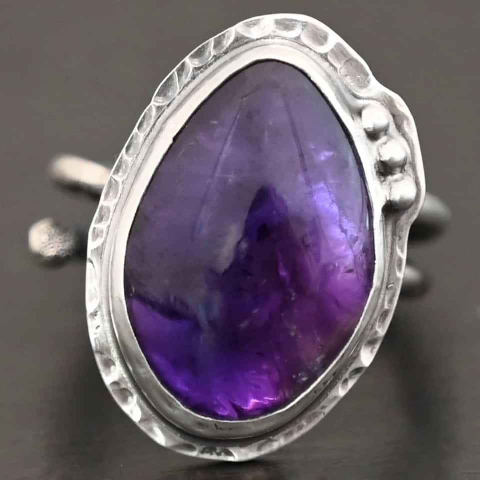 Shades Of Evening Ring Photo of the "Shade's Of Evening" Ring. Made out of sterling silver and amethyst. The amethyst is a slight teardrop shape, but with a rounded tip. The amethyst is a dark purple shade of color, with light purple illuminating parts of the bottom of it. The ring is set in silver. Going around the ring setting is hammered sterling silver. On the bottom left of the amethyst are three silver balls. They are right next to one another, and go in a curve to follow the edge of the amethyst. Ring has one band that loops around to create a double band that can be adjusted to any size. Ring is sitting against a dark background.
