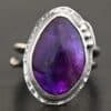 Photo of the "Shade's Of Evening" Ring. Made out of sterling silver and amethyst. The amethyst is a slight teardrop shape, but with a rounded tip. The amethyst is a dark purple shade of color, with light purple illuminating parts of the bottom of it. The ring is set in silver. Going around the ring setting is hammered sterling silver. On the bottom left of the amethyst are three silver balls. They are right next to one another, and go in a curve to follow the edge of the amethyst. Ring has one band that loops around to create a double band that can be adjusted to any size. Ring is sitting against a dark background.