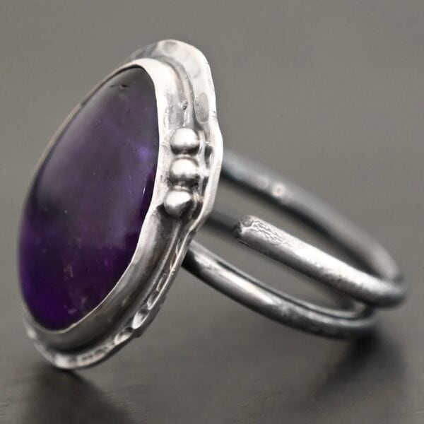 Photo of the "Shade's Of Evening" Ring. Made out of sterling silver and amethyst. The amethyst is a slight teardrop shape, but with a rounded tip. The amethyst is a dark purple shade of color, with light purple illuminating parts of the bottom of it. The ring is set in silver. Going around the ring setting is hammered sterling silver. On the bottom left of the amethyst are three silver balls. They are right next to one another, and go in a curve to follow the edge of the amethyst. Ring has one band that loops around to create a double band that can be adjusted to any size. Ring is sitting against a dark background.