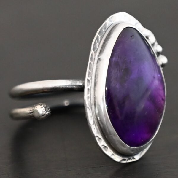 Photo of the "Shade's Of Evening" Ring. Made out of sterling silver and amethyst. The amethyst is a slight teardrop shape, but with a rounded tip. The amethyst is a dark purple shade of color, with light purple illuminating parts of the bottom of it. The ring is set in silver. Going around the ring setting is hammered sterling silver. On the bottom left of the amethyst are three silver balls. They are right next to one another, and go in a curve to follow the edge of the amethyst. Ring has one band that loops around to create a double band that can be adjusted to any size. Ring is sitting against a dark background.